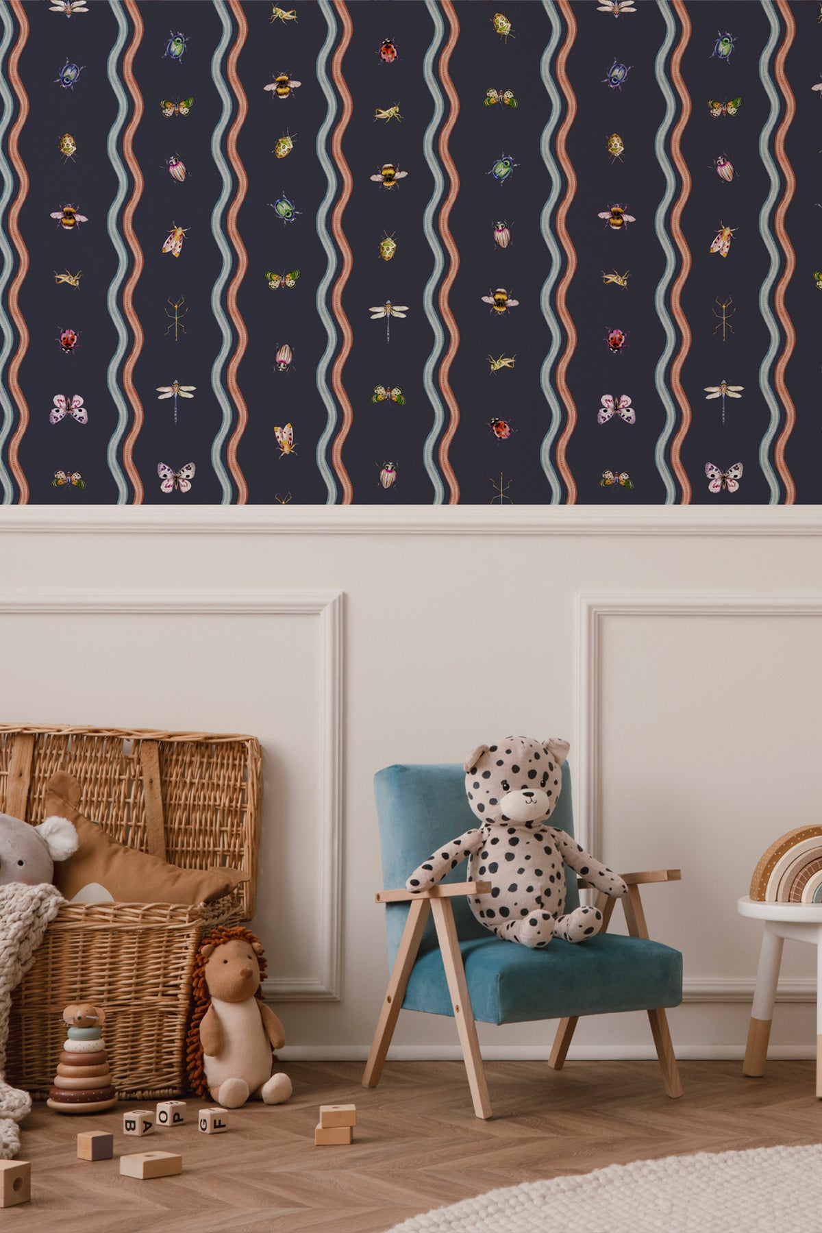 A styled children's room corner showing a navy wallpaper with vertical wavy stripes and small colorful insect motifs above a teal armchair with a spotted teddy bear, plus a wicker basket and toys on a wood floor.