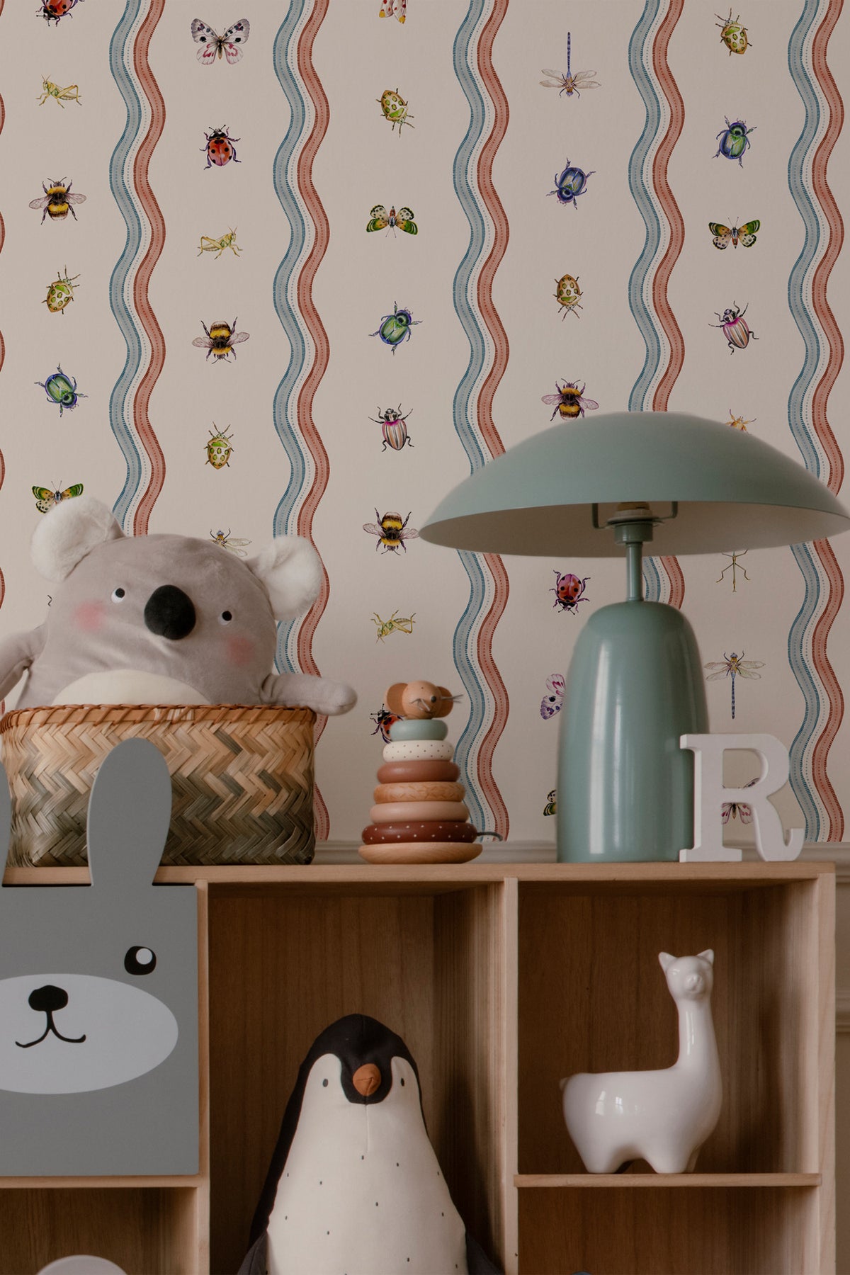 A styled children's room corner featuring patterned wallpaper with wavy vertical stripes and small insect motifs, a wooden shelf with toys, a mint green table lamp, a plush koala in a basket, and decorative animal figurines.