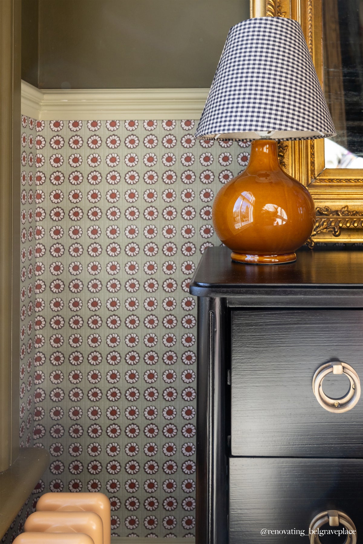 A close-up lifestyle shot of a dark wood dresser with a glossy amber ceramic lamp and a blue-and-white gingham lampshade, set against a wall with patterned wallpaper.