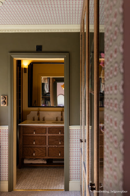 An interior architectural view looking through a doorway into a vintage-style bathroom with a wooden vanity, patterned wallpaper, and tiled walls.