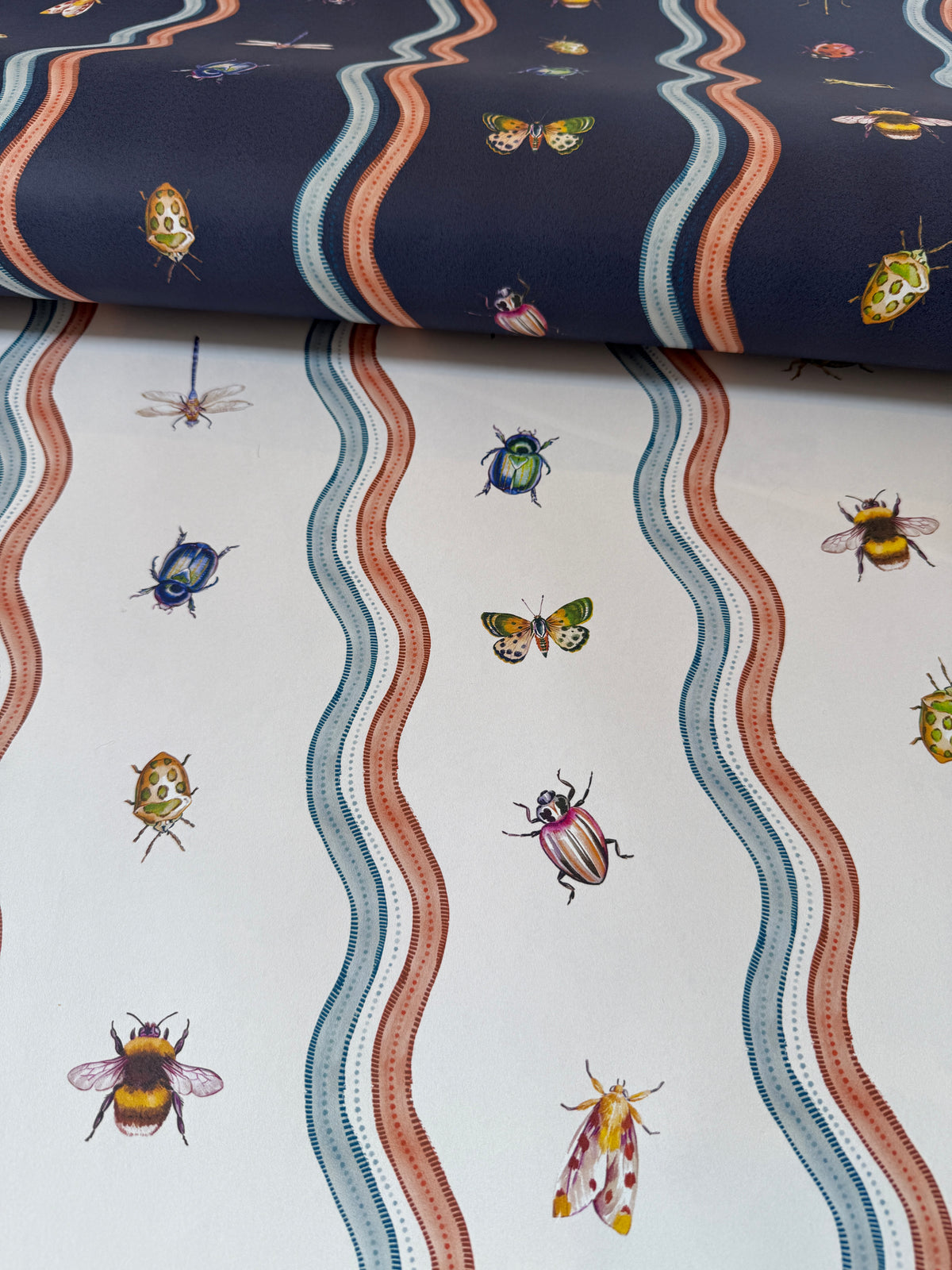 A close-up of patterned fabric or wallpaper showing two sections: one navy blue with wavy coral, cream, and blue stripes and small insects, and one white with matching wavy stripes and illustrated bugs including bees, butterflies, beetles, and moths.