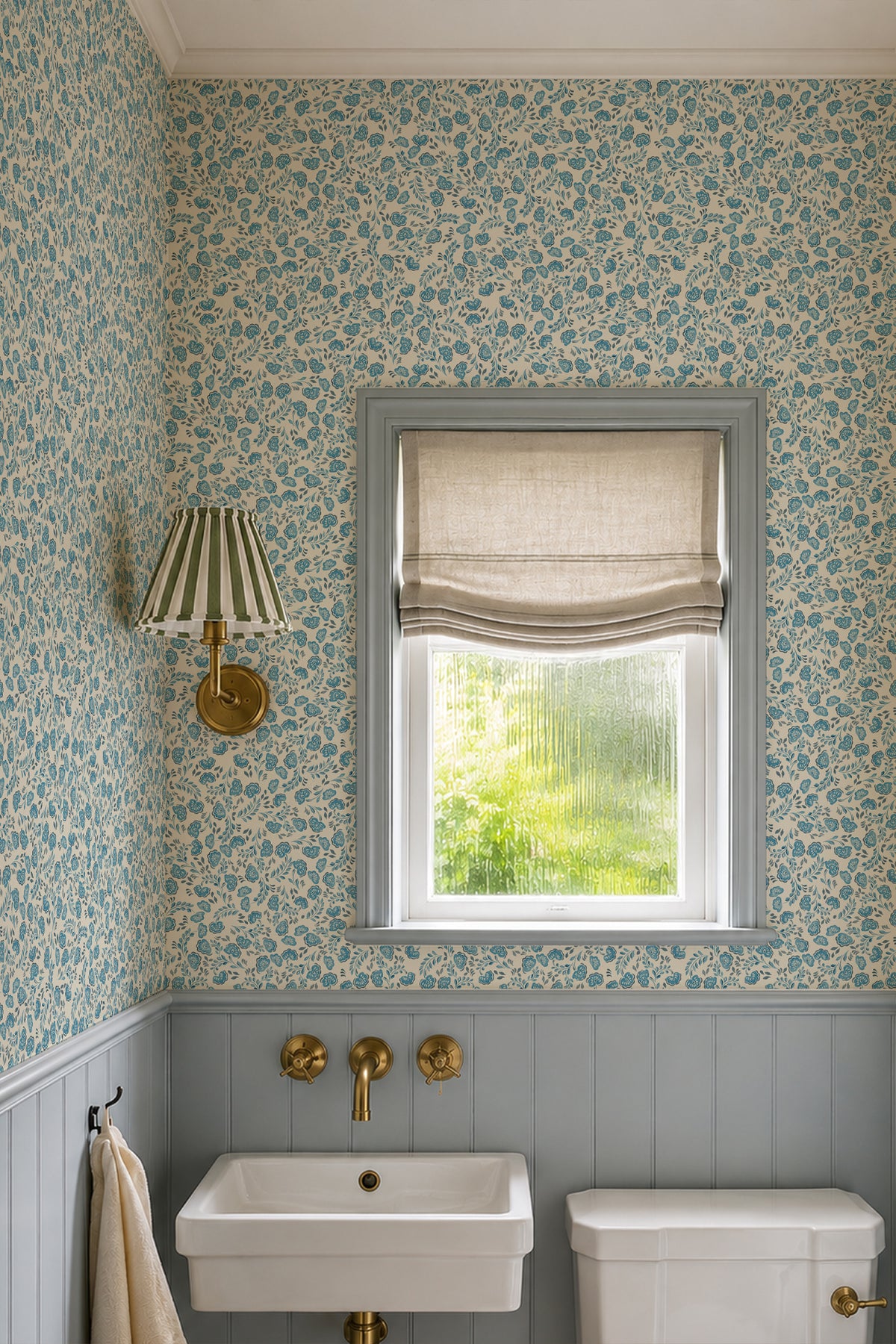 A styled bathroom corner featuring blue and cream floral patterned wallpaper, a gray window frame with a beige roller blind, a white wall-mounted sink, and brass fixtures.