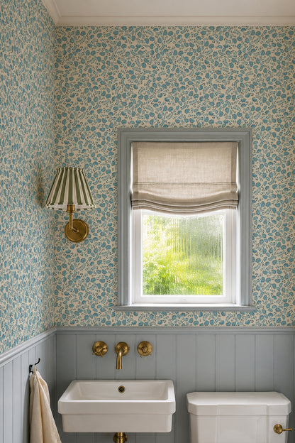 A styled bathroom corner featuring blue and cream floral patterned wallpaper, a gray window frame with a beige roller blind, a white wall-mounted sink, and brass fixtures.