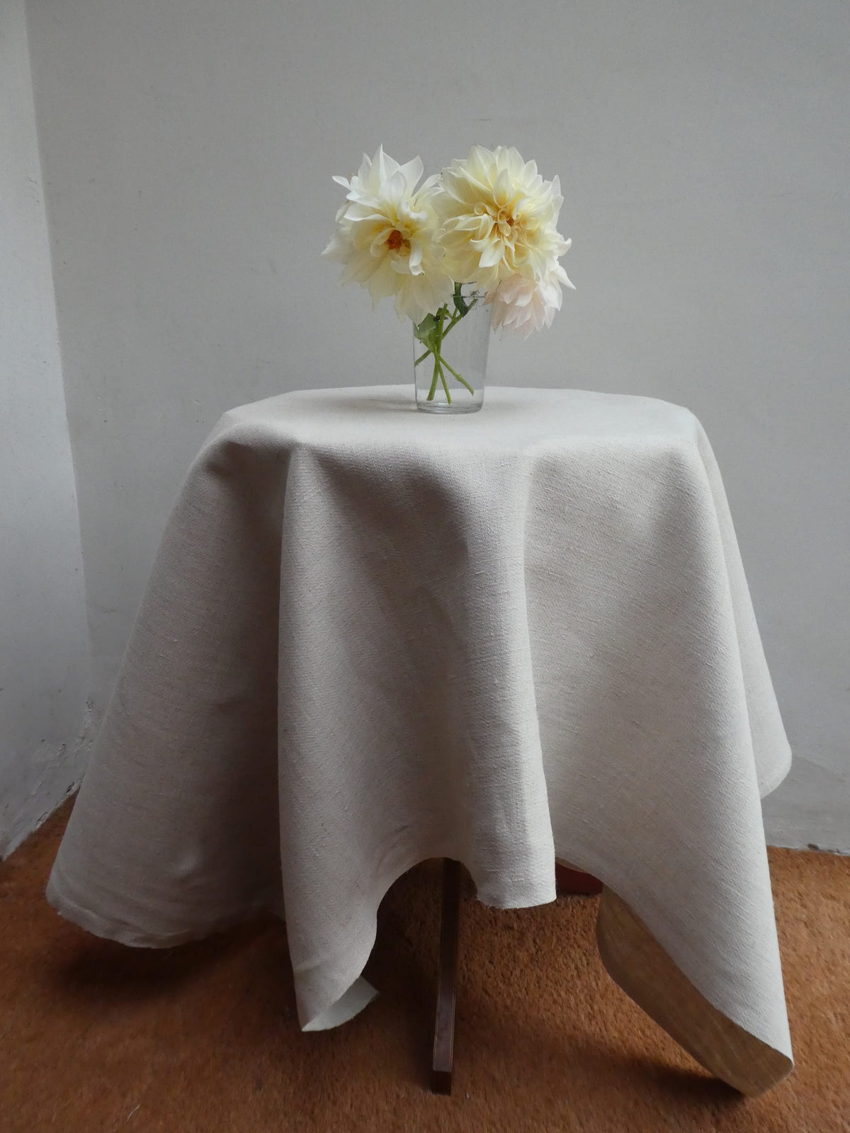 White tablecloth on a small round table with a vase of white flowers against a plain wall.
