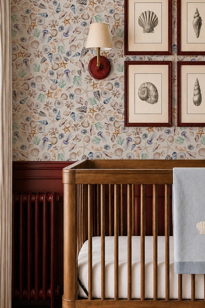 A styled nursery corner featuring a wooden crib with a light blue blanket, a red wall sconce, and a wall covered in nautical seashell and starfish wallpaper. Above the crib, four framed seashell illustrations are mounted on the wall.