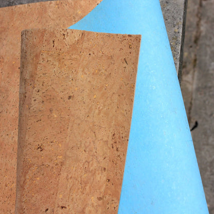 A close-up of a cork sheet with a natural tan-brown textured surface, partially peeled back to reveal a bright light blue backing, set against a concrete surface.
