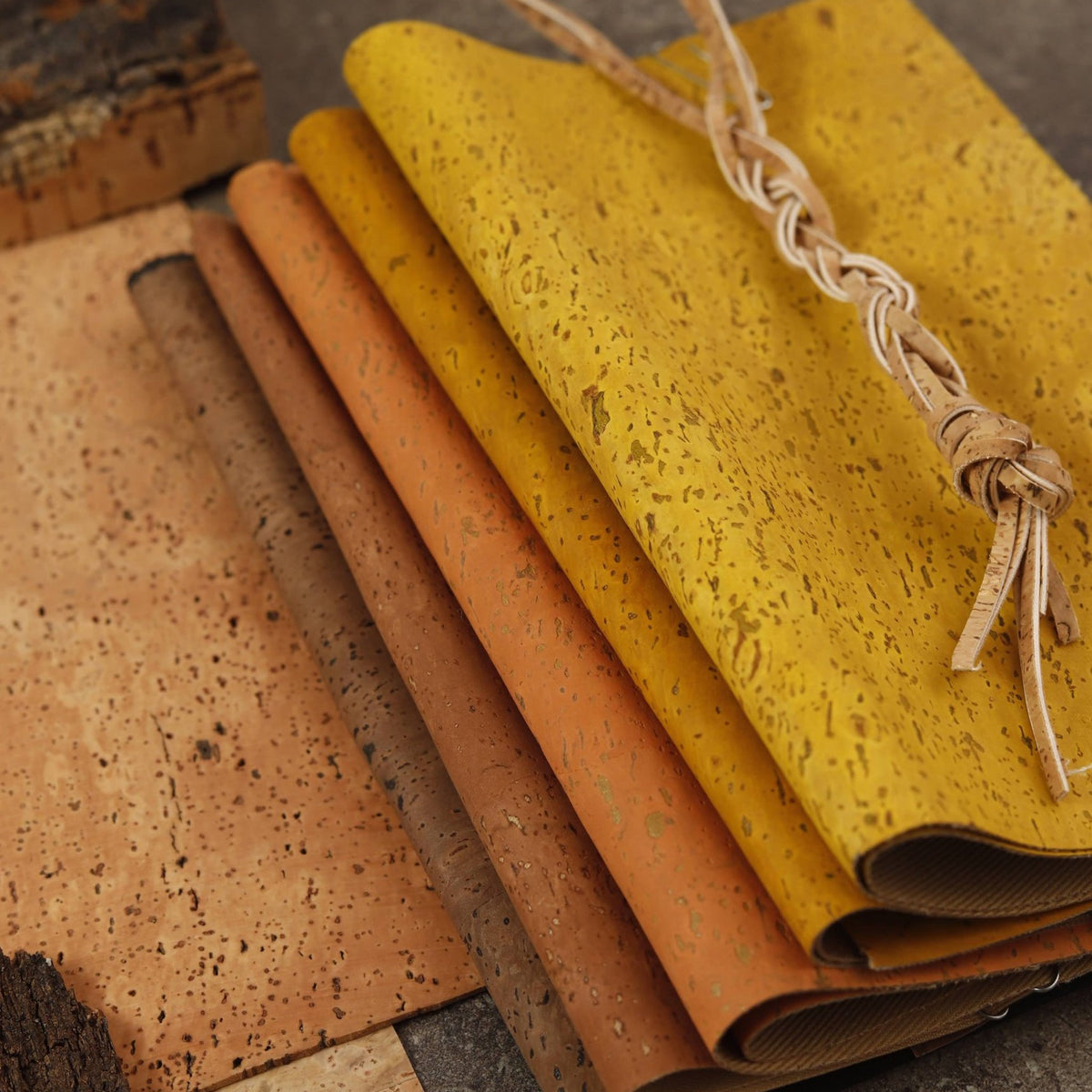 A stack of cork sheets in various earthy tones including mustard yellow, terracotta orange, brown, and tan, with a braided twine handle resting on top, set against a dark textured background.