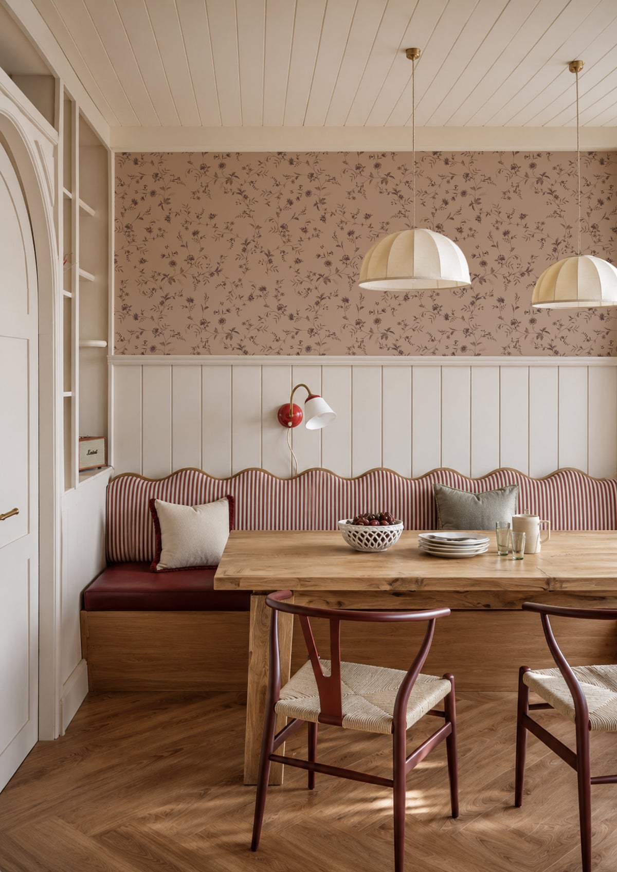 A cozy dining nook with a wooden table, two chairs, a red upholstered bench, and a floral wallpapered accent wall in a bright, sunlit room.