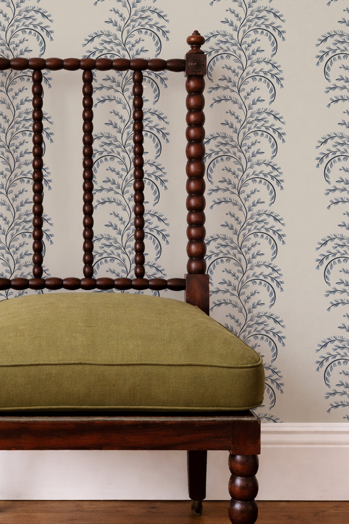 A close-up of a dark brown wooden spindle-back chair with an olive green cushion, set against a wall with blue and white damask wallpaper.