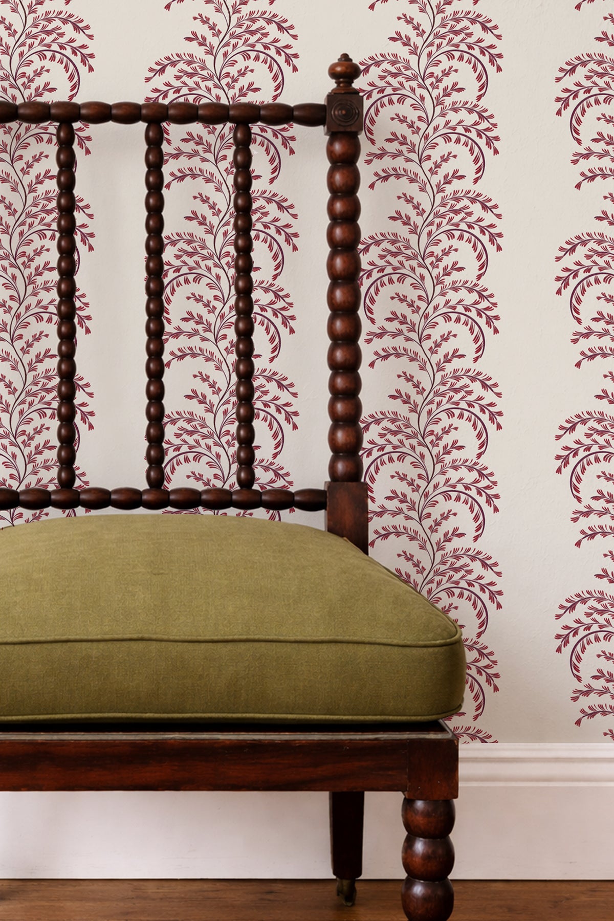 A close-up of a dark wood spindle-back chair with an olive green cushion, set against a wall with cream wallpaper featuring a burgundy floral vine pattern.