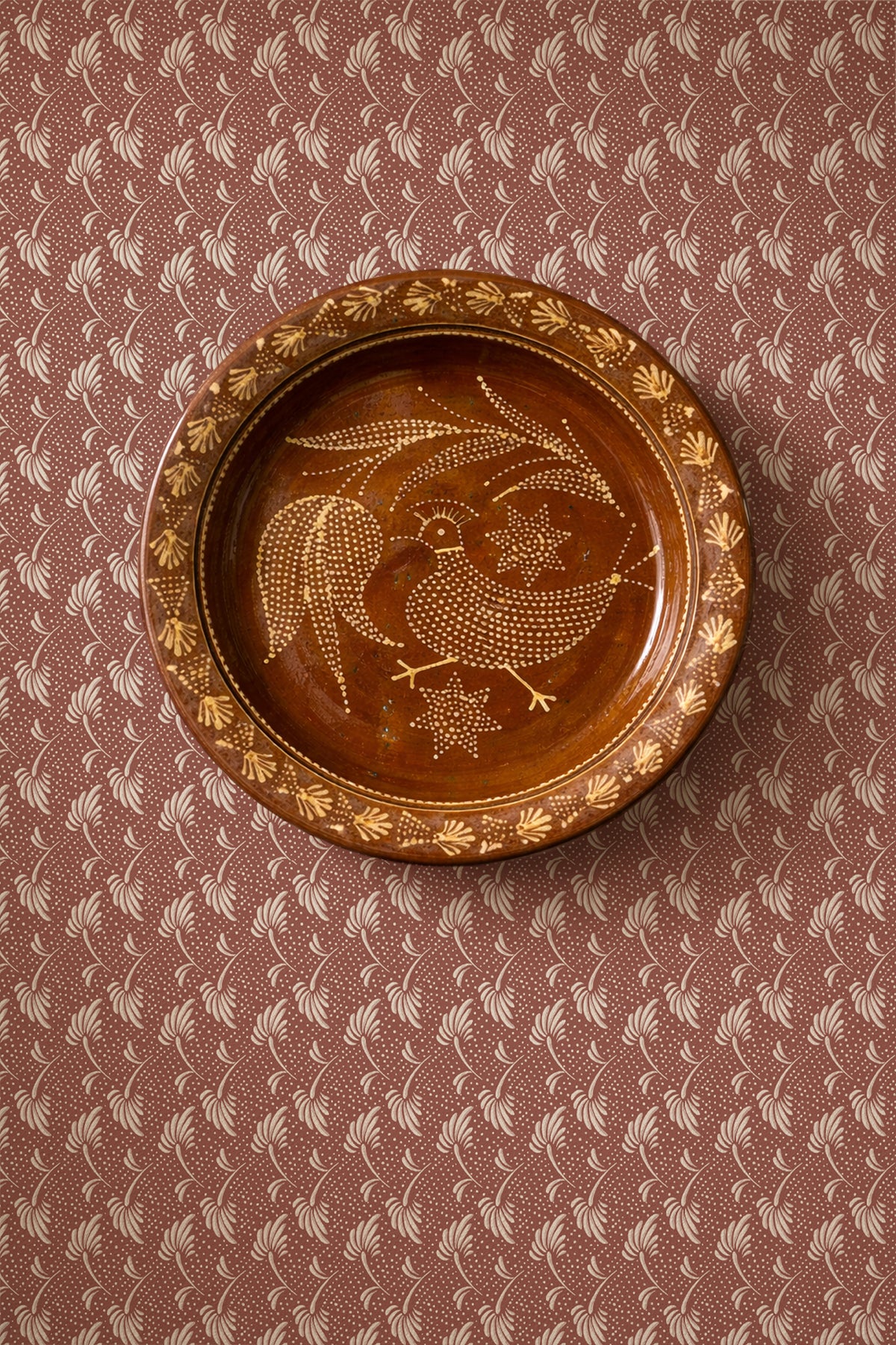 A round brown ceramic plate with a hand-painted bird and floral design, displayed on a patterned brown and beige surface.