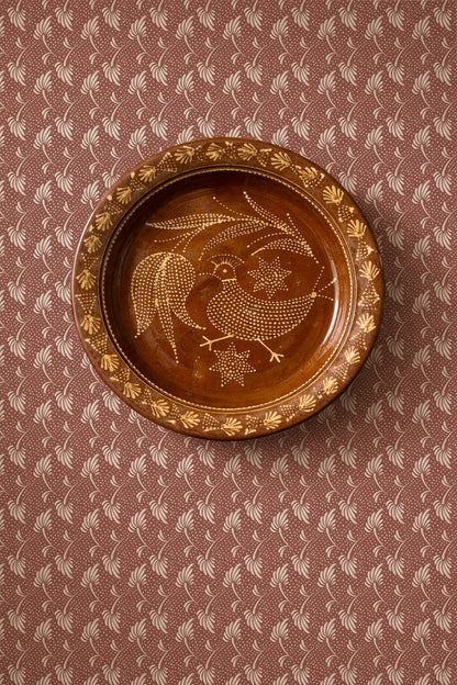 A round brown ceramic plate with a hand-painted bird and floral design, displayed on a patterned brown and beige surface.