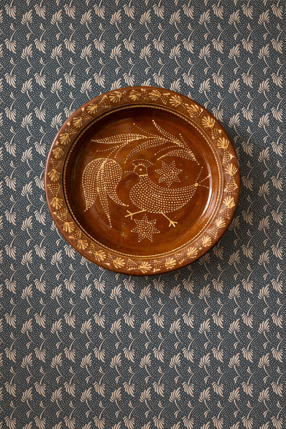 A round brown ceramic plate with hand-painted gold and white floral and bird motifs, displayed on a dark patterned fabric background.