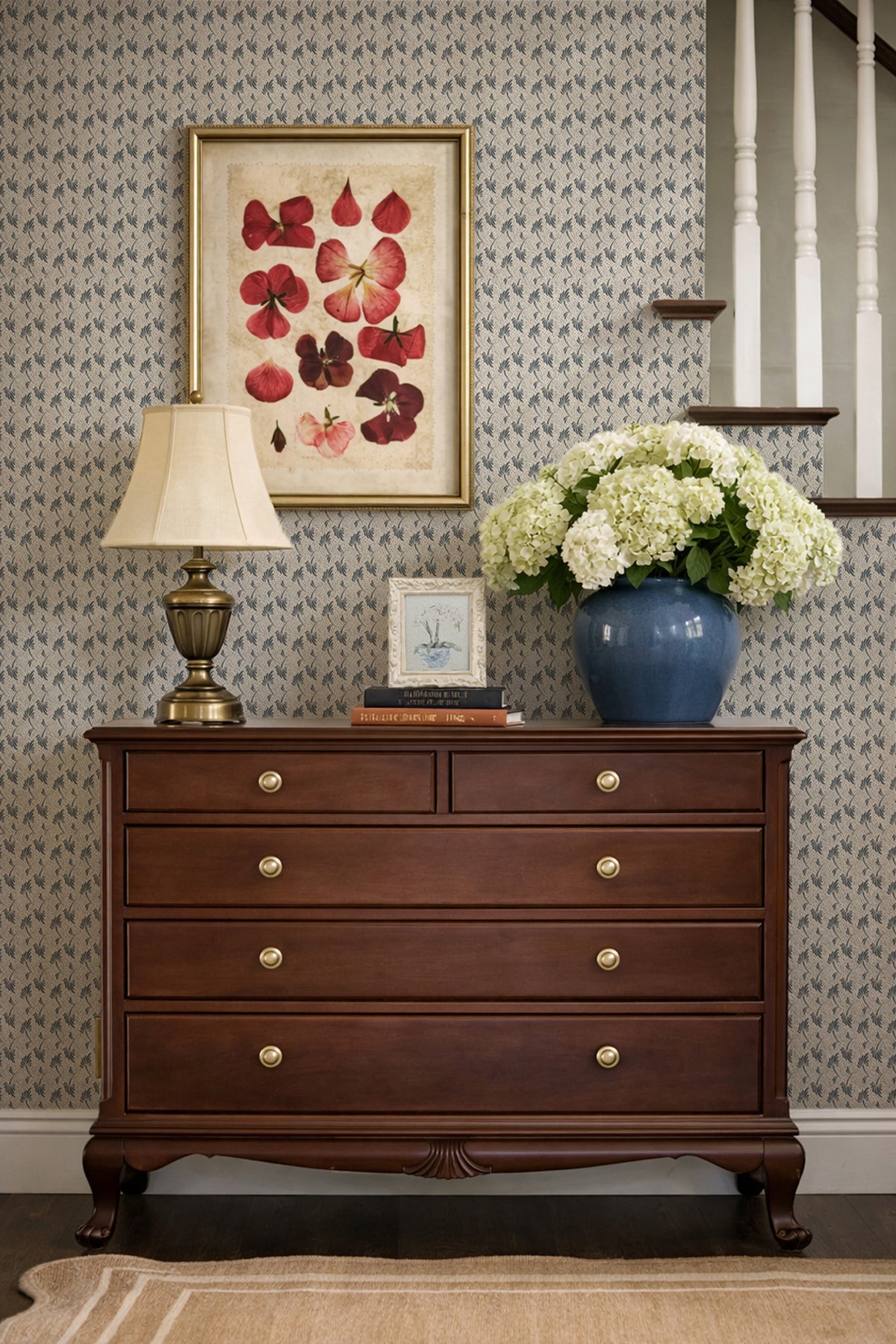 A dark brown wooden chest of drawers with brass knobs, styled in an entryway against patterned wallpaper. A table lamp, a framed floral print, and a blue vase with white hydrangeas sit on top.