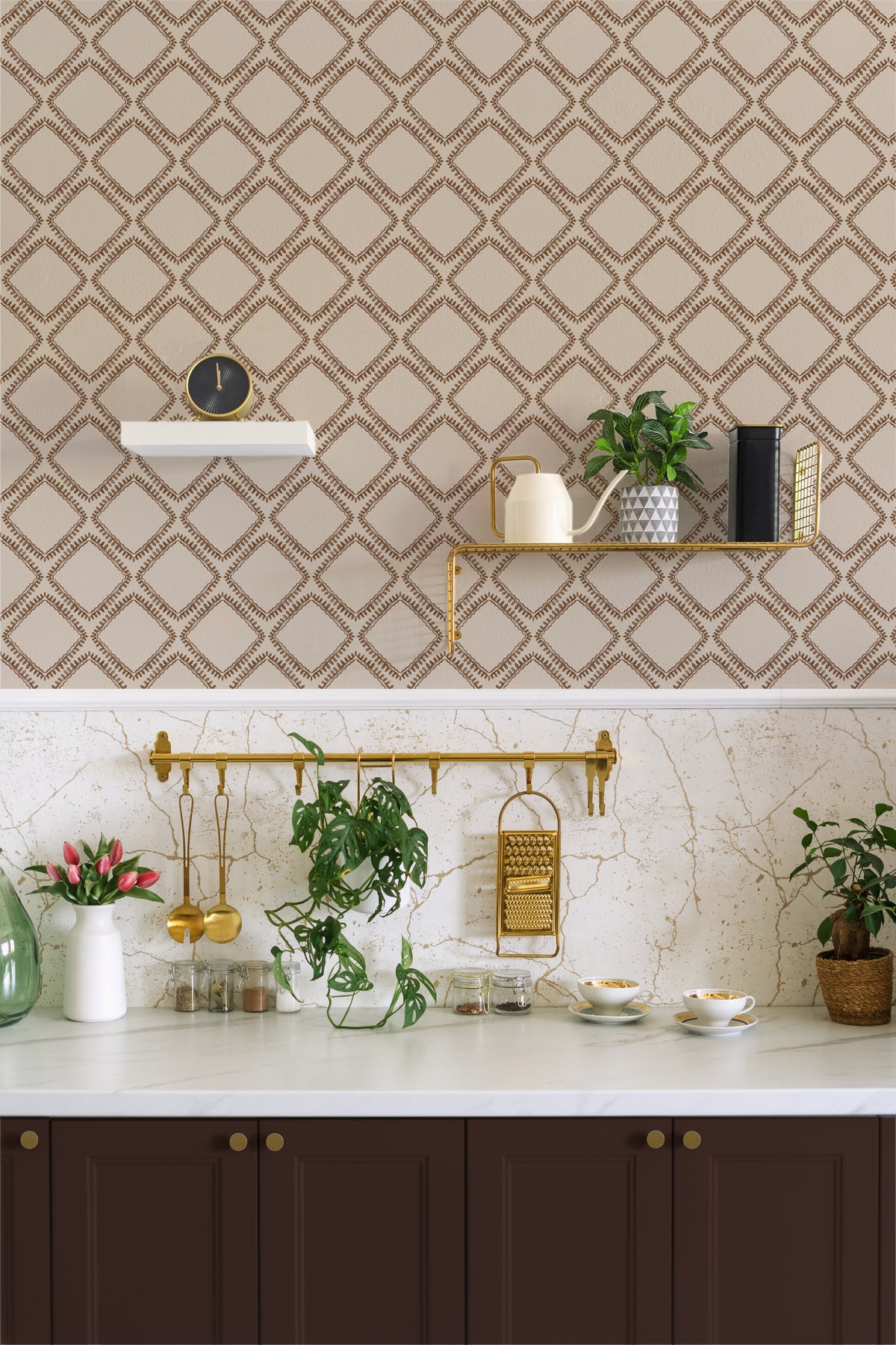 A styled kitchen interior featuring beige wallpaper with a brown geometric trellis pattern on the upper wall, a white marble countertop, dark brown cabinets, gold wall hardware, and several potted green plants.