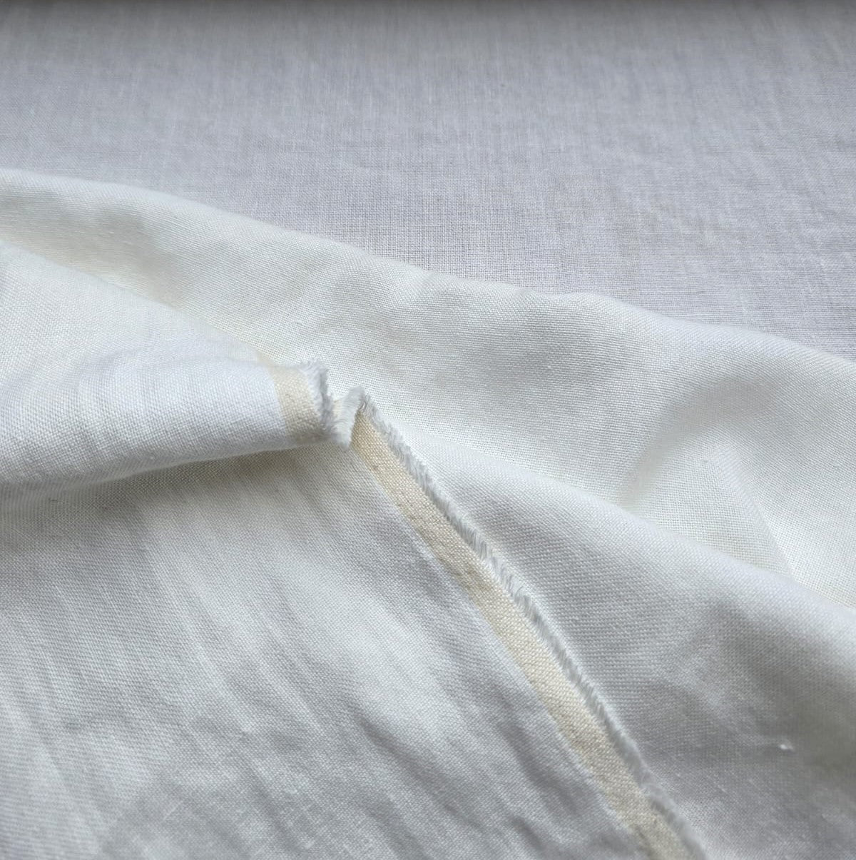 A close-up of off-white fabric with a visible fold and raw, slightly frayed edge, showing a light tan underside.