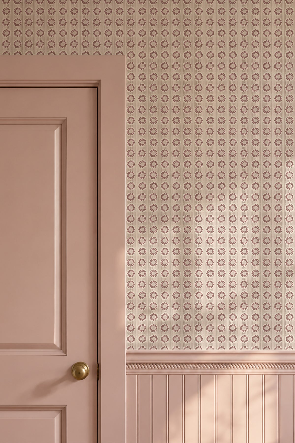 A close-up of a dusty pink interior wall with a repeating circular geometric pattern on the upper section and wainscoting below, alongside a matching pink paneled door with a brass doorknob.