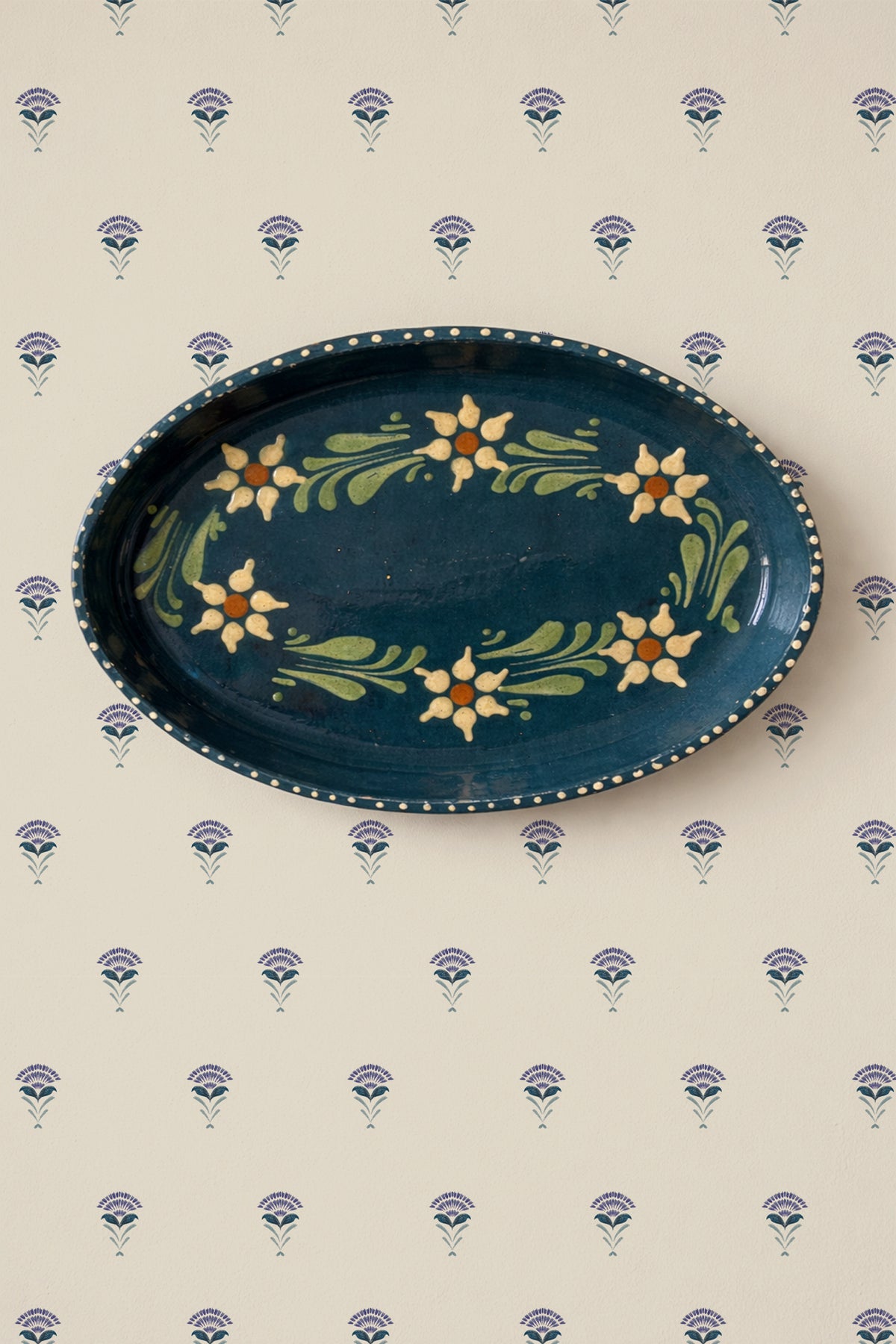 An oval dark blue ceramic serving platter with a hand-painted floral border, displayed on a cream wallpaper with a repeating blue and purple floral pattern.