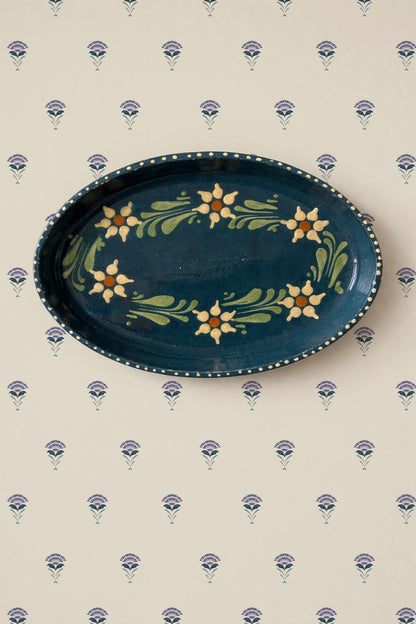 An oval dark blue ceramic serving platter with a hand-painted floral border, displayed on a cream wallpaper with a repeating blue and purple floral pattern.