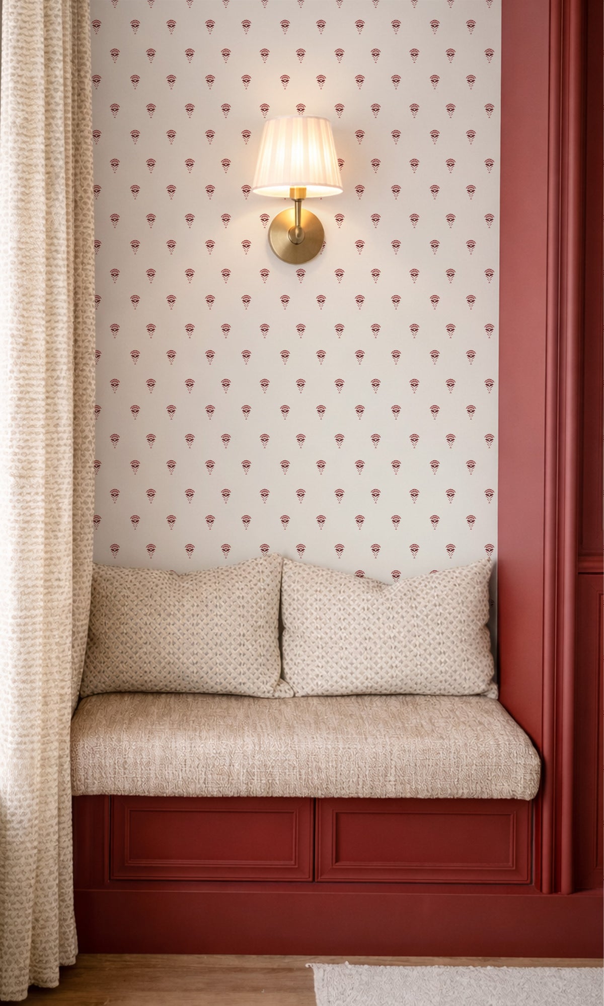 A cozy built-in window seat with a textured beige cushion and two matching pillows, set against a wall with red trim and a repeating floral wallpaper pattern. A warm brass wall sconce with a cream shade is mounted above the seat.