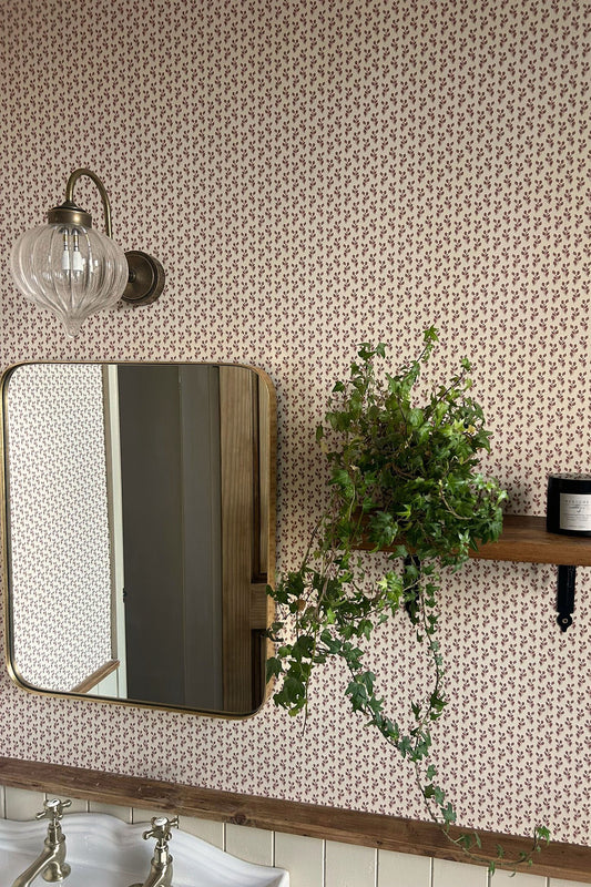 A bathroom scene with patterned wallpaper, a wall-mounted mirror, a glass wall sconce, a wooden shelf holding a green trailing plant, and part of a white sink visible at the bottom.