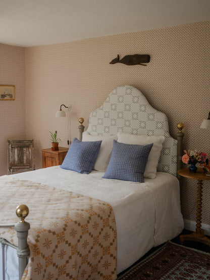 A styled bedroom featuring a bed with a patterned cream and gray upholstered headboard, white bedding, two blue checkered pillows, and a beige floral-patterned quilt. The room has pink patterned wallpaper, wooden bedside tables, and a dark wood fish wall decor.