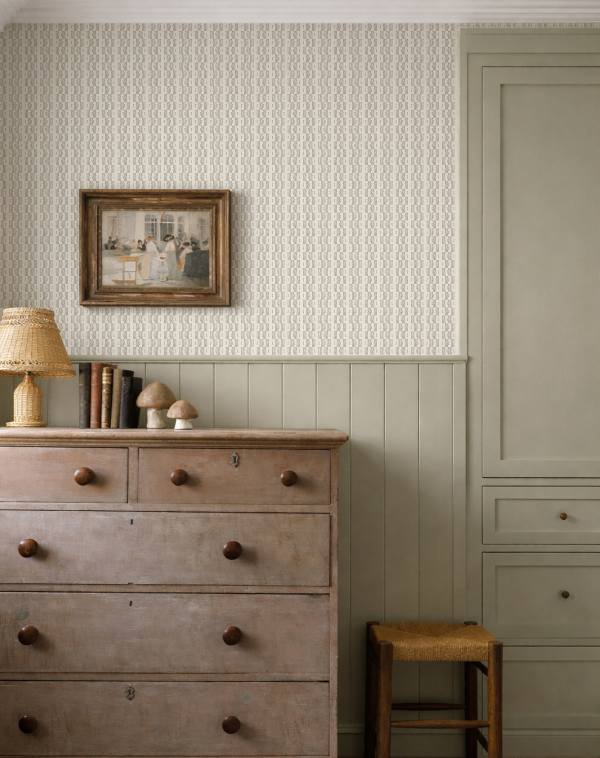 A styled interior room scene showing wallpaper on the upper wall, a light wood dresser with dark knobs, a small lamp, framed artwork, wainscoting, and a muted green cabinet.