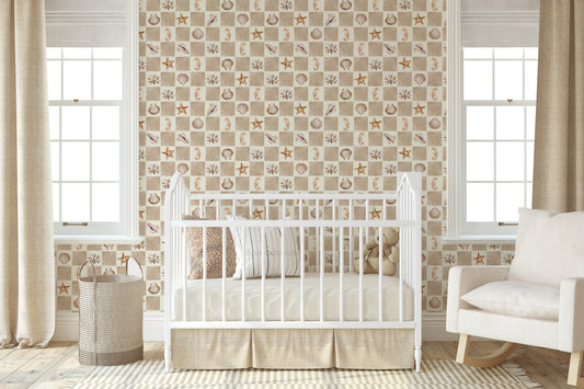 A styled nursery room featuring a white wooden crib with a ruffled skirt, set against a wall with nautical-themed wallpaper. The wallpaper has a beige and white checkered pattern with illustrations of seashells, starfish, and small boats.