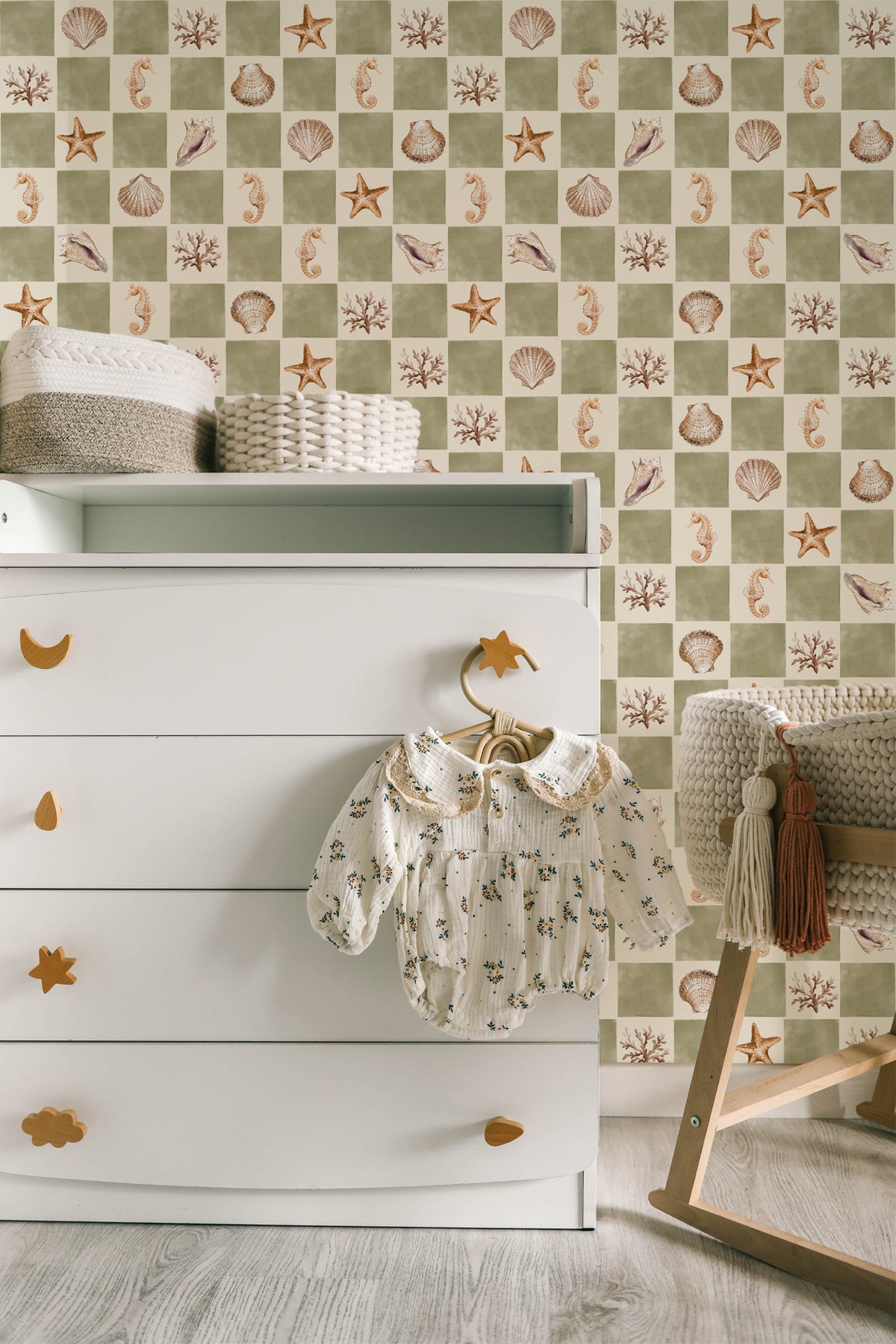 A styled nursery corner featuring a white wooden dresser with gold moon and star-shaped pulls, a baby romper hanging on a horse-shaped hanger, and a patterned wallpaper in the background with sea urchins, starfish, and shells in beige and sage green.