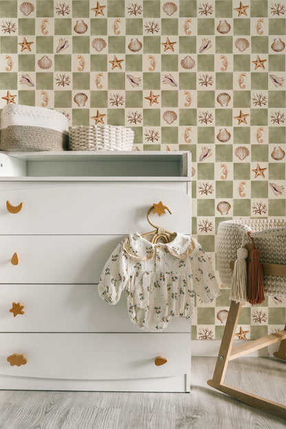 A styled nursery corner featuring a white wooden dresser with gold moon and star-shaped pulls, a baby romper hanging on a horse-shaped hanger, and a patterned wallpaper in the background with sea urchins, starfish, and shells in beige and sage green.