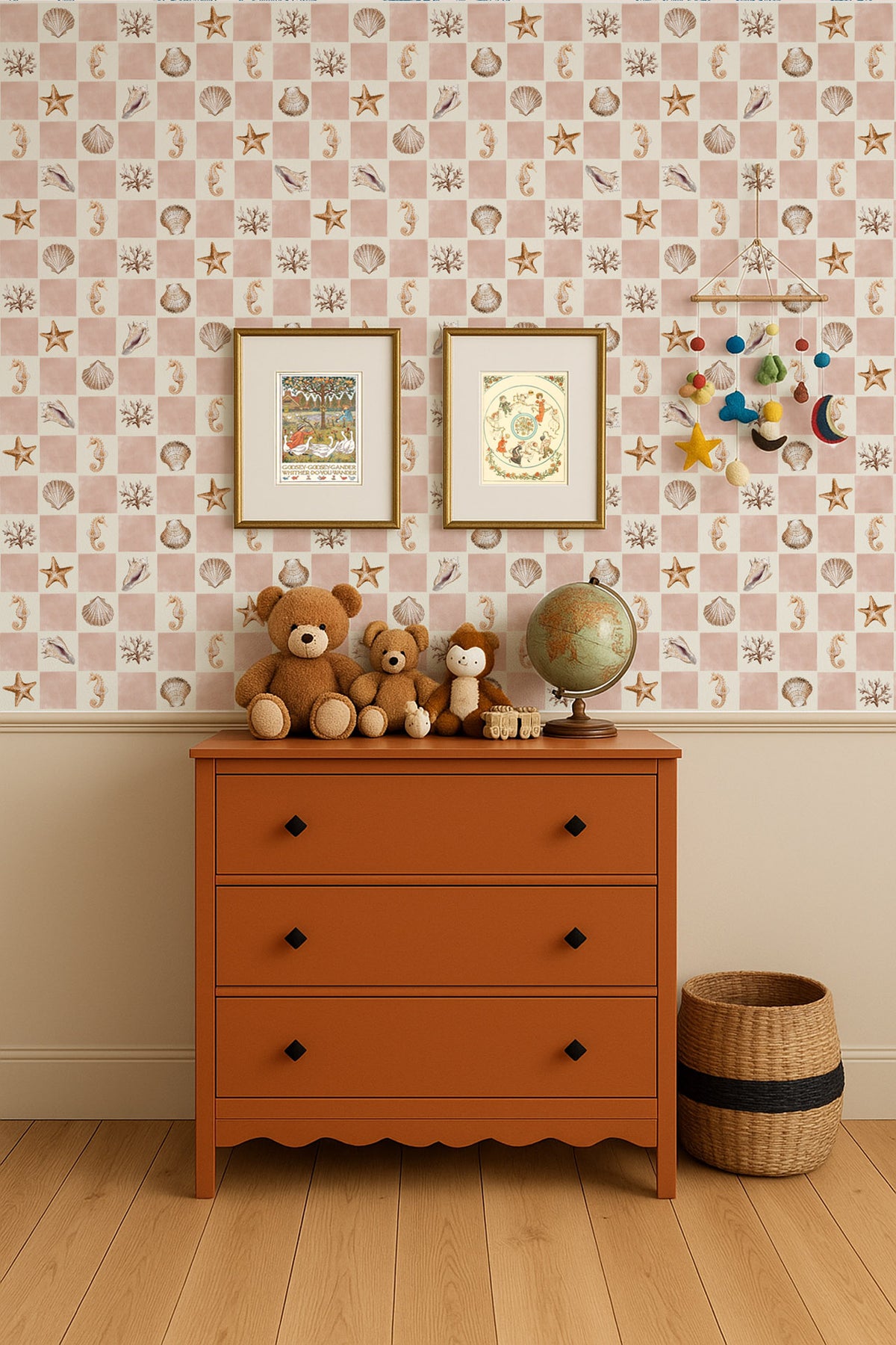 A styled children's room corner featuring a terracotta orange three-drawer dresser against a pink and white checkered wallpaper with sea-themed motifs. The dresser is decorated with three brown teddy bears, a vintage-style globe, and small wooden figurines, with two framed prints hanging on the wall above.