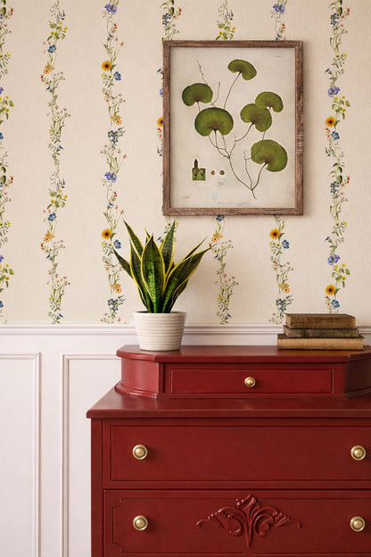 A styled interior scene featuring a red wooden chest of drawers with brass knobs, a potted snake plant, and a framed botanical print on floral wallpaper.