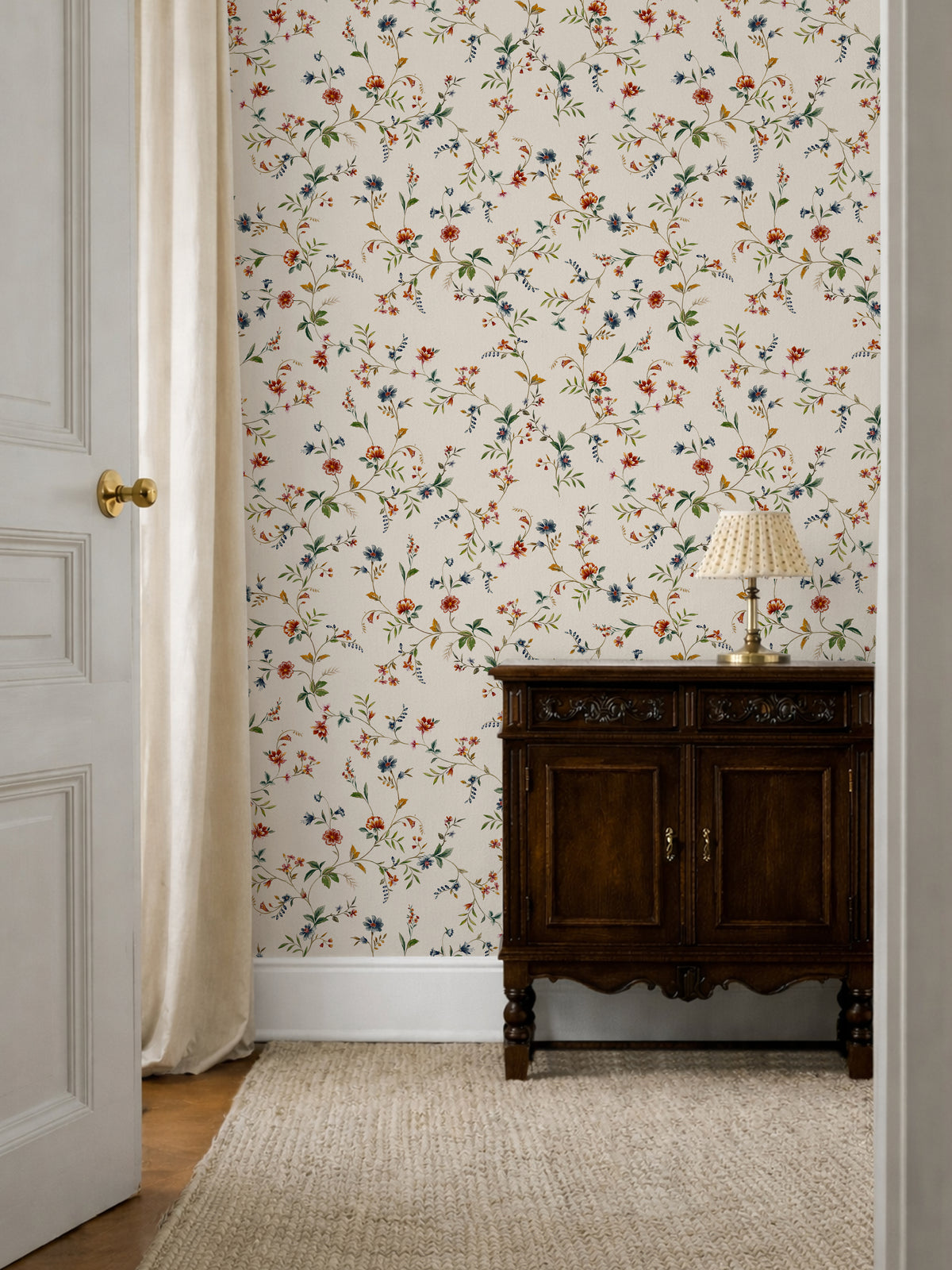 A styled interior room corner featuring floral wallpaper with small orange, blue, and green flowers on a cream background, a dark wood cabinet, a table lamp, a woven rug, and part of an open white door with a curtain.
