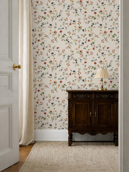 A styled interior room corner featuring floral wallpaper with small orange, blue, and green flowers on a cream background, a dark wood cabinet, a table lamp, a woven rug, and part of an open white door with a curtain.