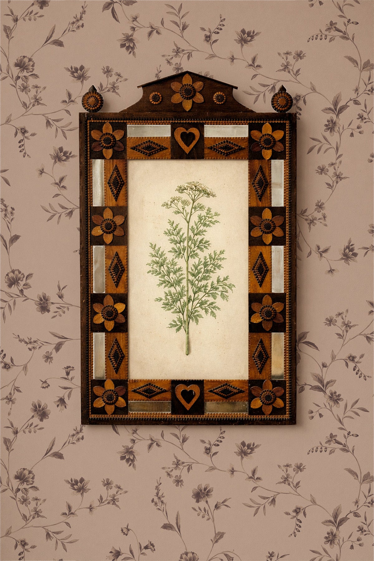 A framed botanical art print featuring a green leafy plant on an off-white background, surrounded by an ornate dark brown and gold geometric border, mounted on a beige floral wallpaper.