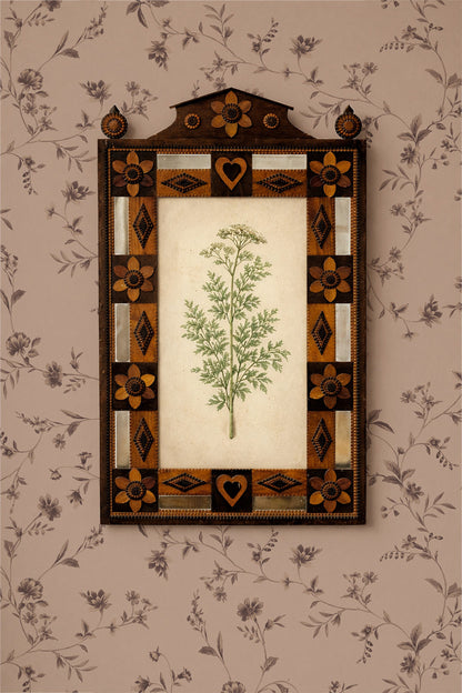 A framed botanical art print featuring a green leafy plant on an off-white background, surrounded by an ornate dark brown and gold geometric border, mounted on a beige floral wallpaper.