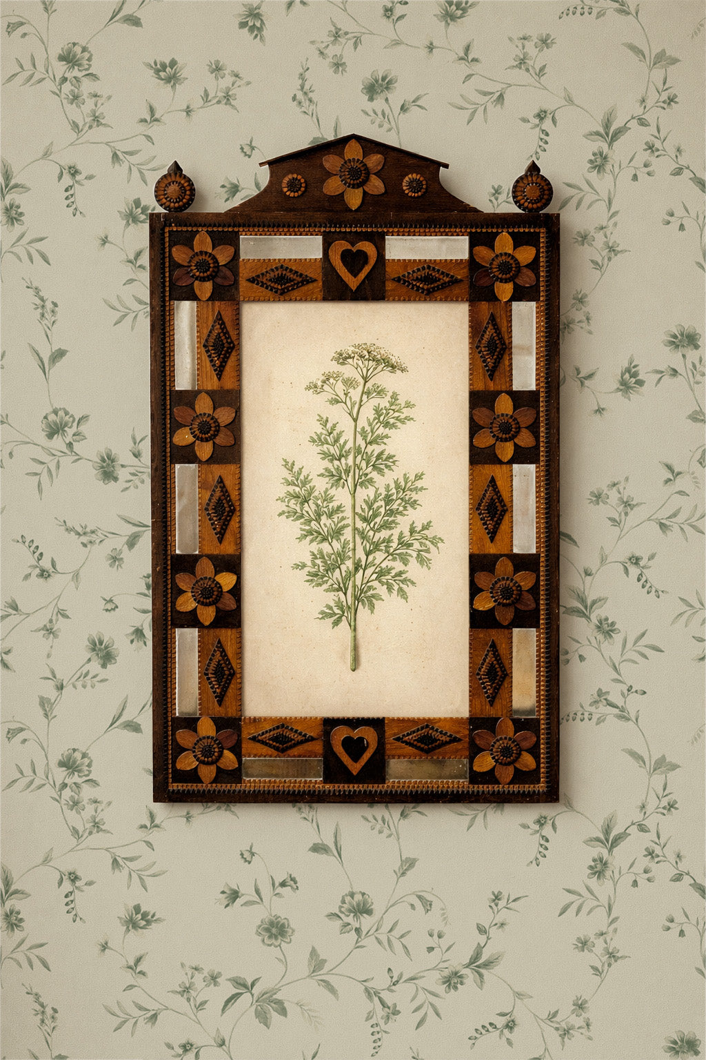 A framed botanical art print featuring a green herb illustration on an off-white background, surrounded by an ornate geometric border in dark brown, tan, and cream. The framed piece is mounted on a light green floral wallpaper.