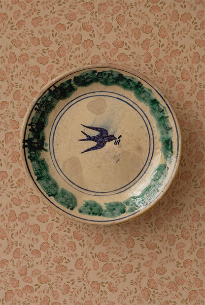 A round ceramic plate with a blue swallow motif in the center and a green painted border, displayed on a pink floral patterned background.