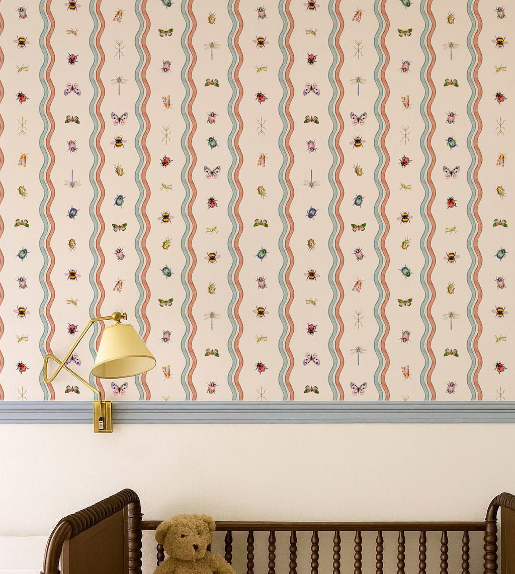 A styled nursery corner showing a wall covered in whimsical vertical wavy stripes with tiny colorful bug and butterfly motifs, above a white wall and above a wooden crib with a teddy bear.