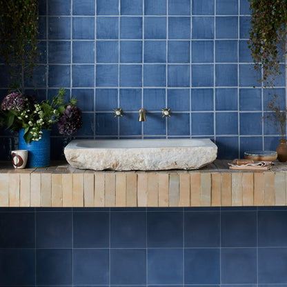 A rustic stone washbasin with a brass faucet set against a blue tiled wall, set on a light wood vanity shelf with decorative flowers and accessories.