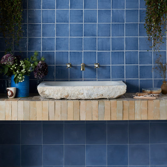A rustic stone washbasin with a brass faucet set against a blue tiled wall, set on a light wood vanity shelf with decorative flowers and accessories.
