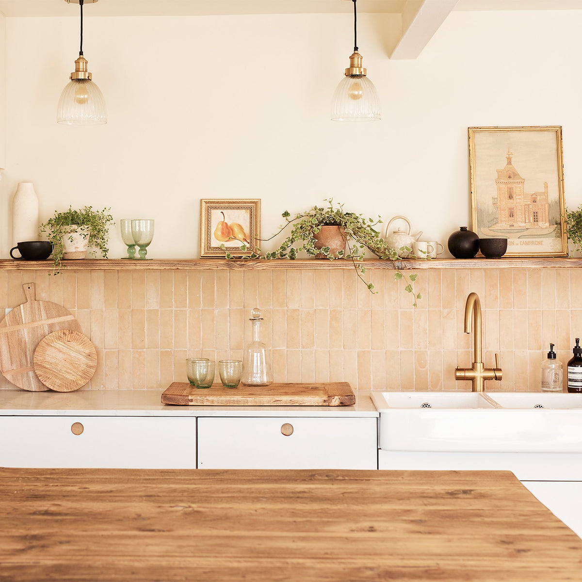 A bright, modern kitchen interior with white cabinets, a beige tile backsplash, a wooden countertop, and a brass faucet. A wooden shelf above the counter holds potted green plants, glassware, and framed wall art.