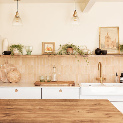 A bright, modern kitchen interior with white cabinets, a beige tile backsplash, a wooden countertop, and a brass faucet. A wooden shelf above the counter holds potted green plants, glassware, and framed wall art.