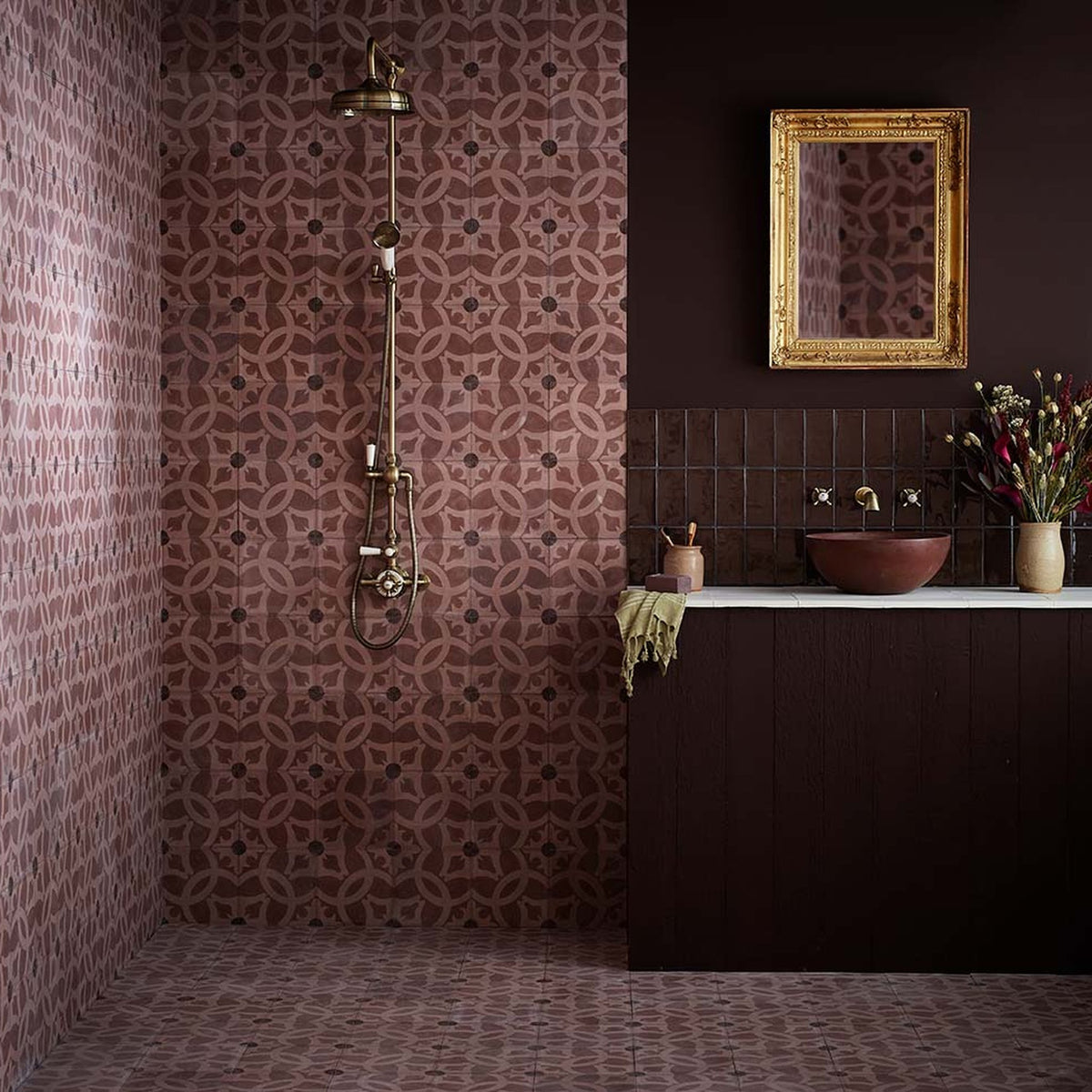 A styled bathroom interior featuring patterned terracotta and brown mosaic tiles, a dark wood vanity with a white countertop, a terracotta vessel sink, and a gold-framed mirror.
