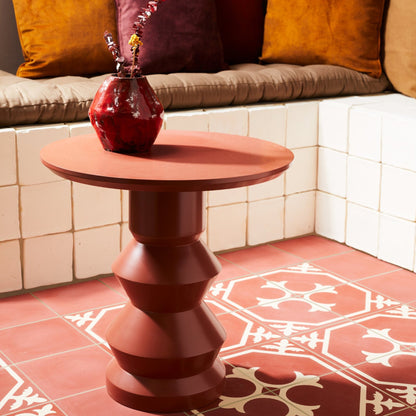 A terracotta-colored round side table with a sculptural, tiered pedestal base, styled in a cozy living room setting with a matching red vase on top.