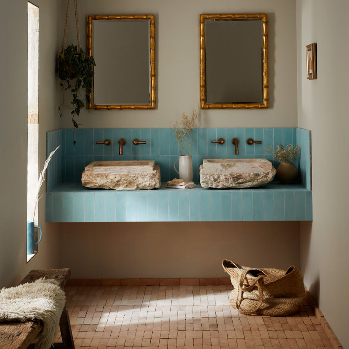 A styled bathroom interior featuring a turquoise tiled double vanity with two stone vessel sinks, two gold-framed mirrors, and warm neutral decor including woven baskets, a bench with white fur, and dried plants.