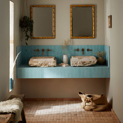 A styled bathroom interior featuring a turquoise tiled double vanity with two stone vessel sinks, two gold-framed mirrors, and warm neutral decor including woven baskets, a bench with white fur, and dried plants.